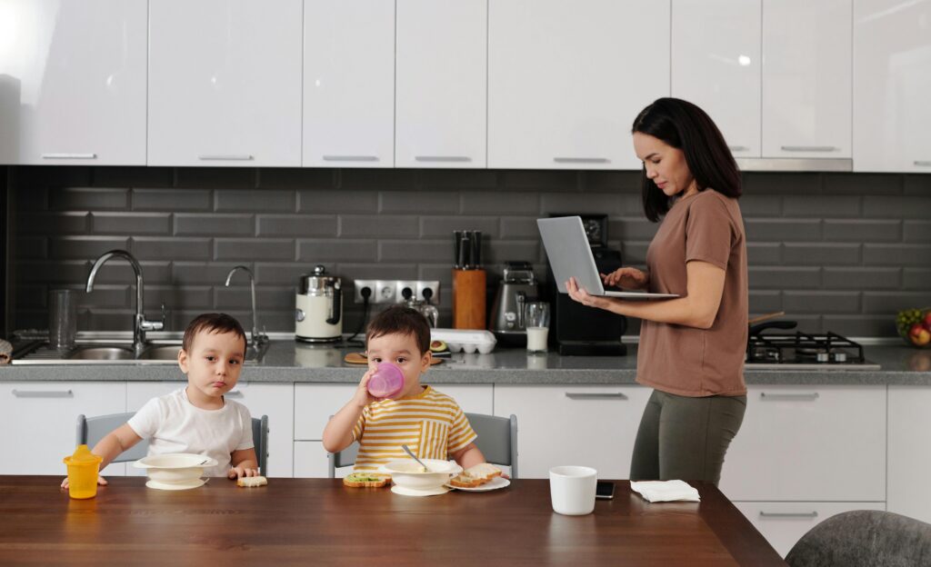 A mother multitasking with a laptop while her children enjoy breakfast at home.